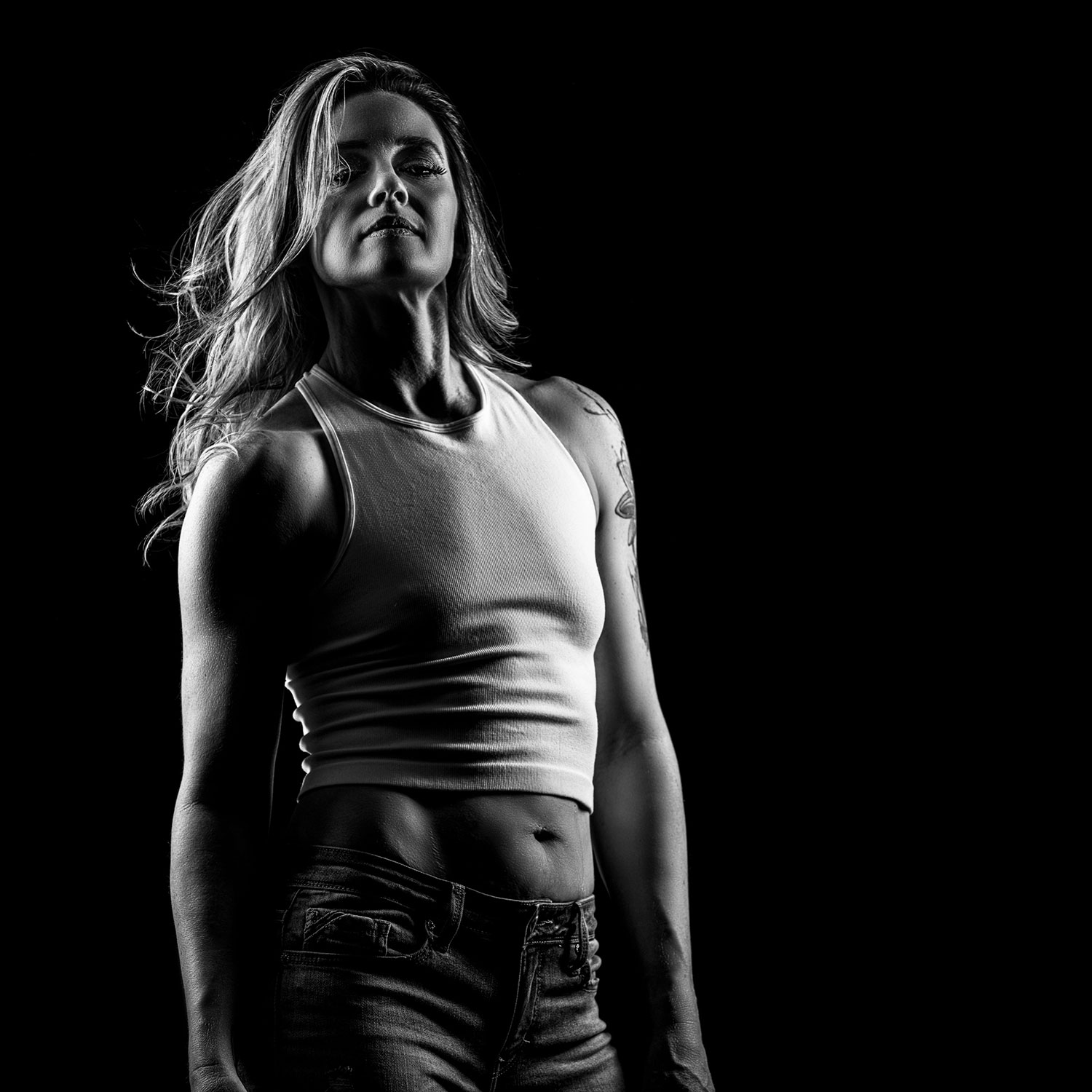 A monochrome image of a fit woman in white tanktop and jeans posing with confidence against a dark background