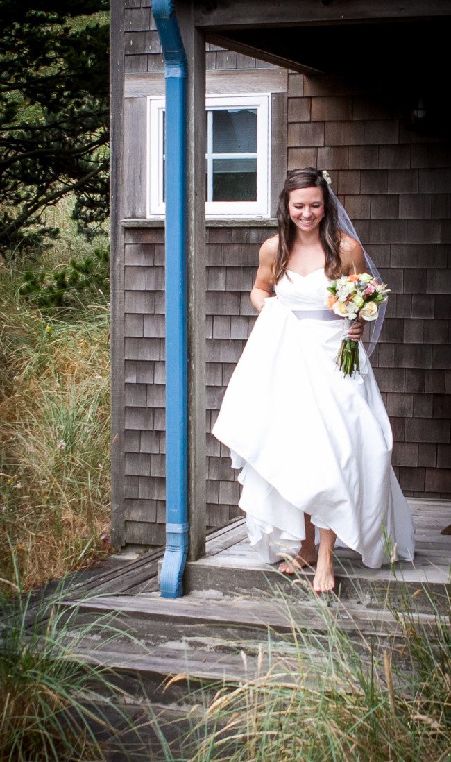 Joyful bride stepping out of a wooden cabin, holding a bouquet, with a natural backdrop, epitomizing elegance and simplicity.