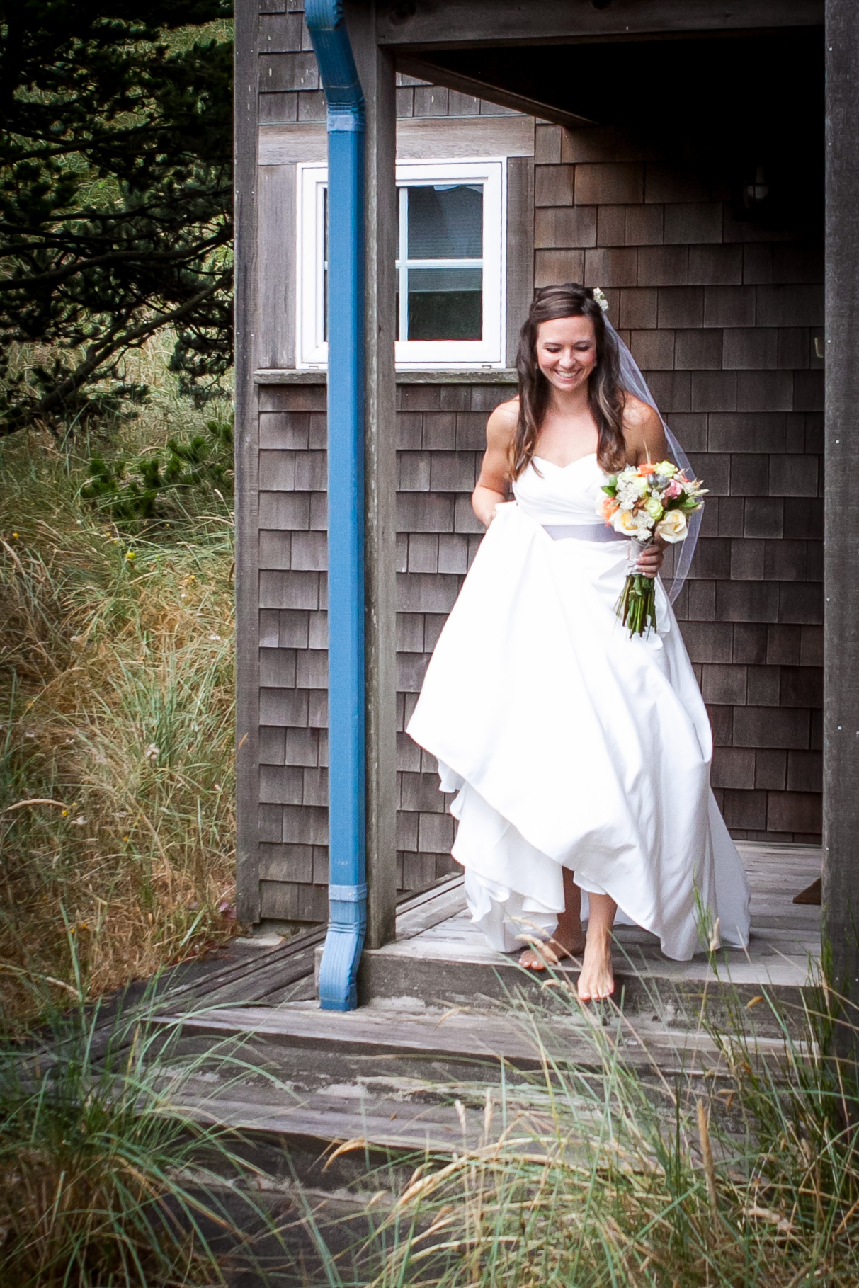 Joyful bride stepping out of a wooden cabin, holding a bouquet, with a natural backdrop, epitomizing elegance and simplicity.