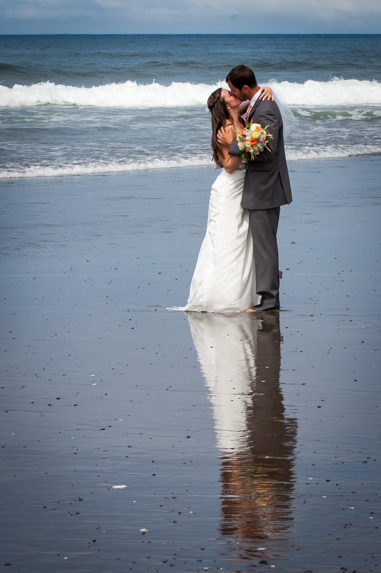 Bride and groom kissing on the beach with ocean waves and reflections on sand