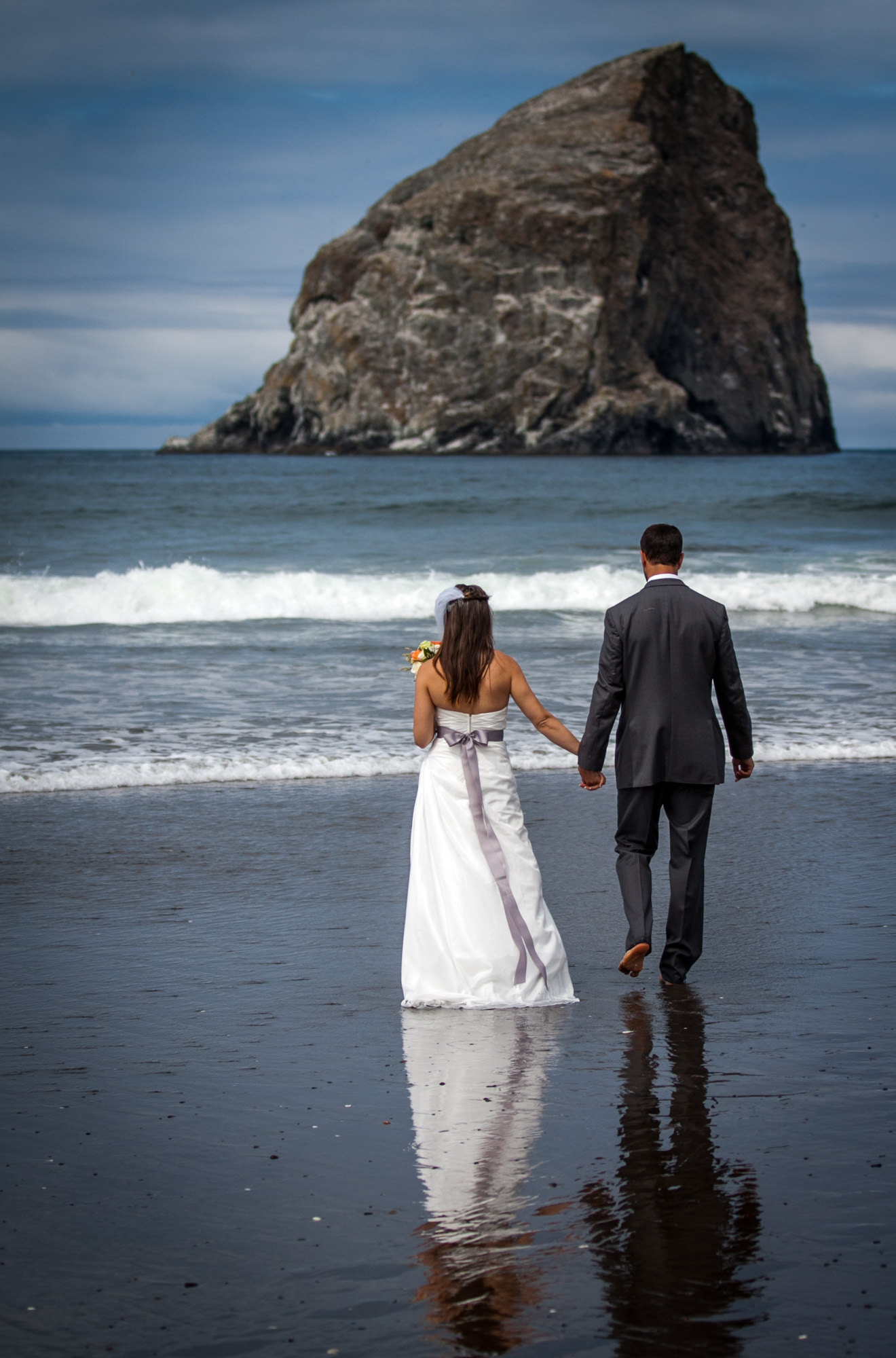 Bride and groom walking hand in hand on the beach, with ocean waves and sea stack in the background, bride carrying a bouquet.