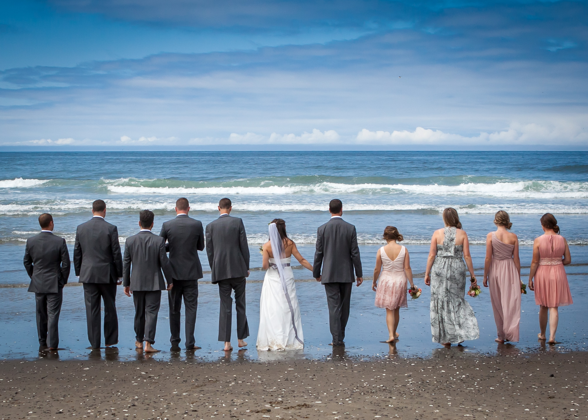 Back view of the wedding party holding hands on the beach, looking out at the ocean.