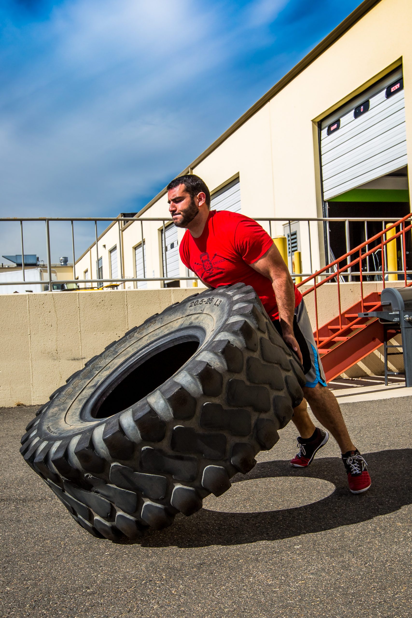 Man in Red Shirt Flipping Large Tractor Tire Outdoors, Bright Blue Sky