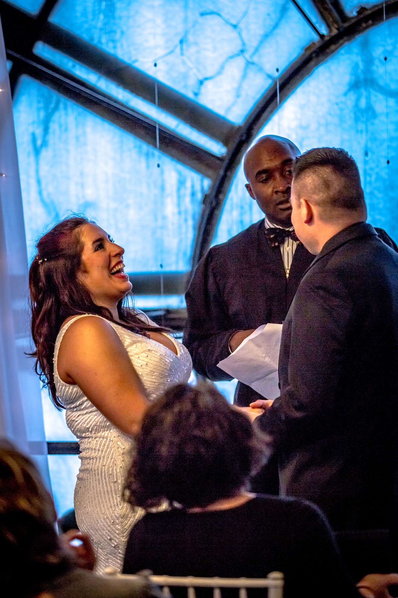 Bride Laughing With Joy During Wedding Ceremony With Officiant and Groom, Under a Striking Circular Window