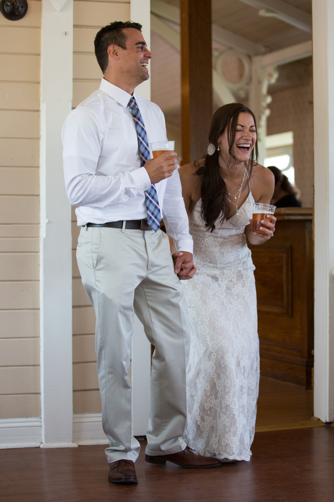 Laughing bride in lace gown with groom in white shirt at wedding reception, both holding drinks