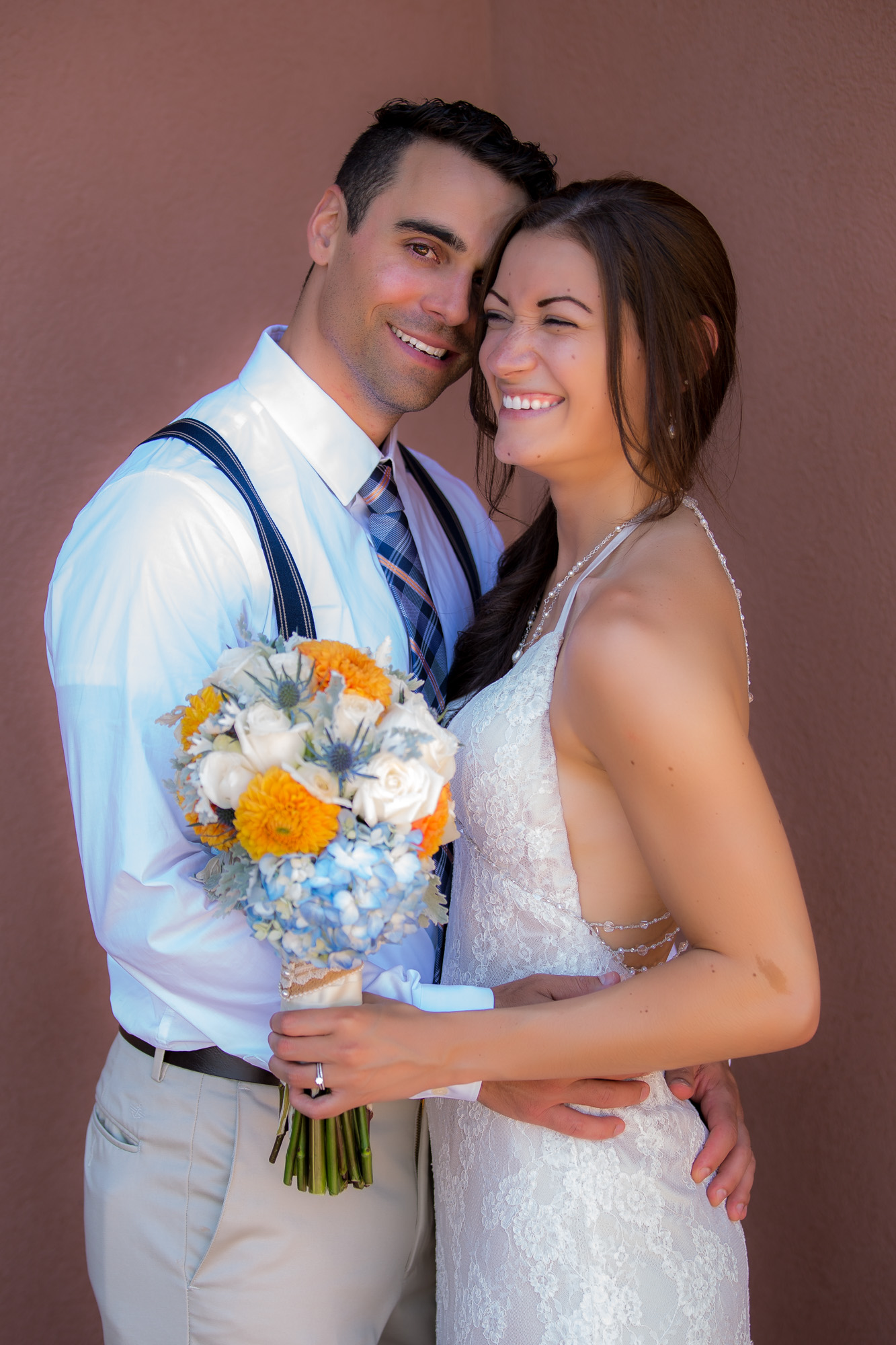 Happy bride in lace gown and groom in suspenders hugging and smiling on their wedding day.