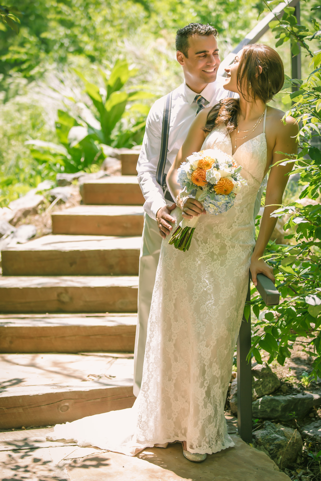 Newlywed couple smiling at each other in nature, bride in lace dress with orange and blue bouquet, groom in beige suit with suspenders