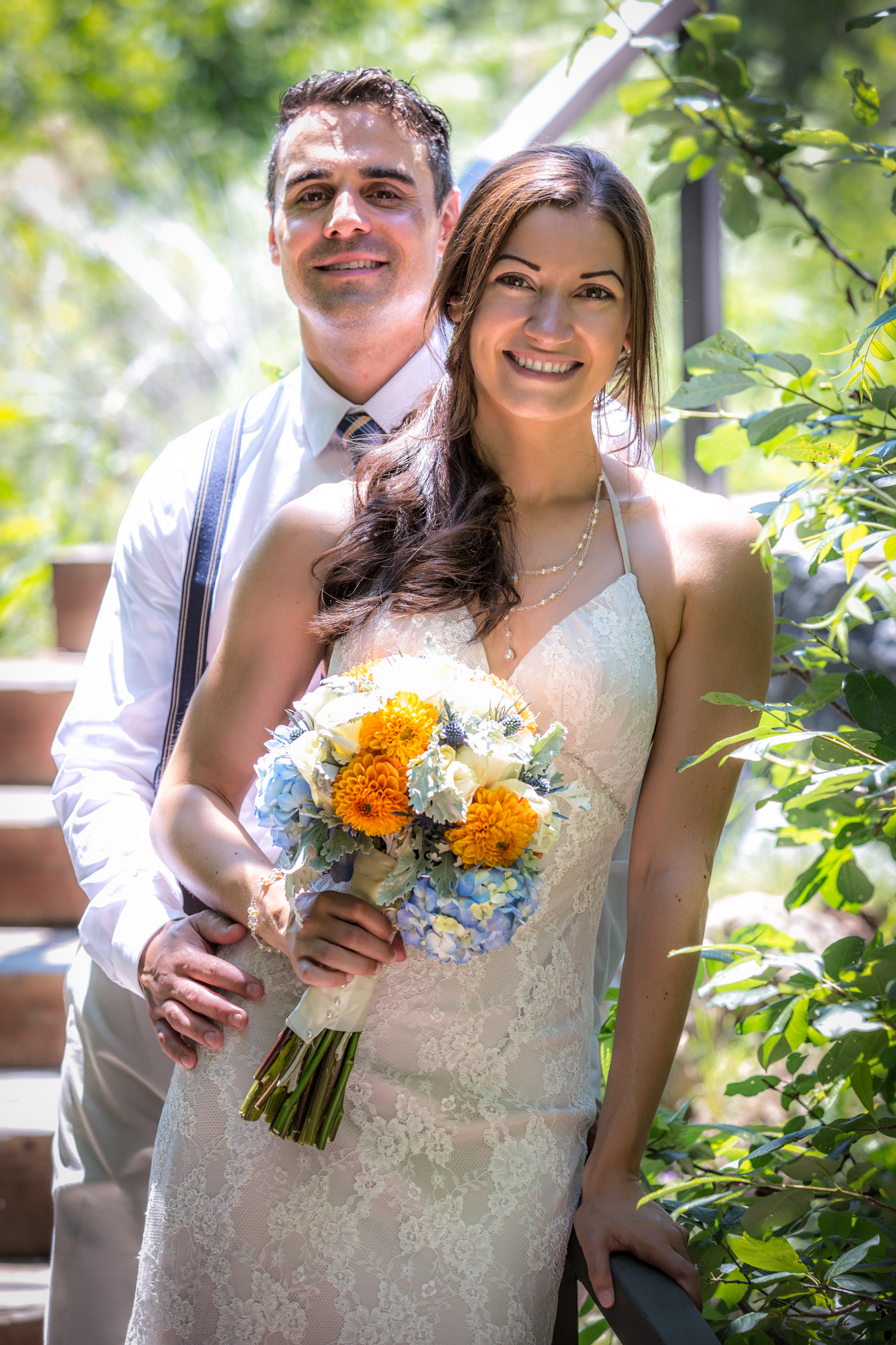 Radiant bride in lace gown with colorful bouquet embraced by groom in suspenders, amidst a natural setting