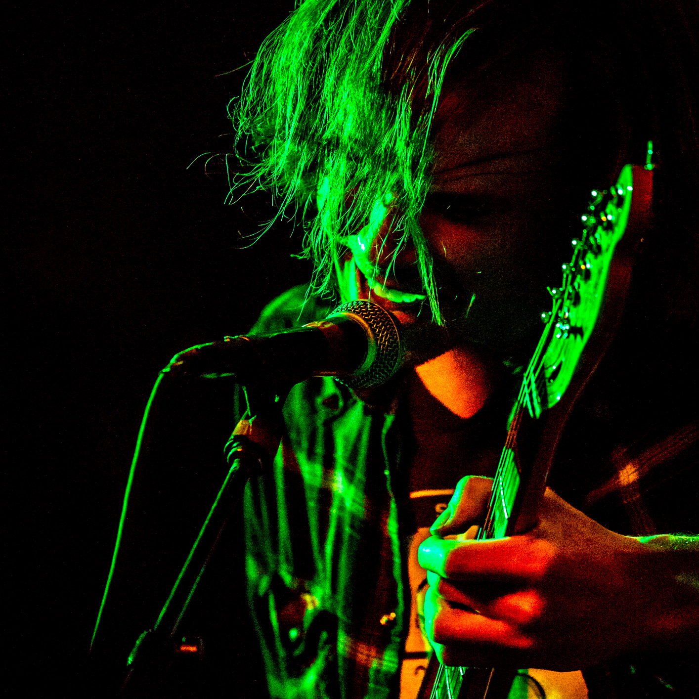 A singer with long hair in his face plays guitar and yells into a microphone during a live musical performance.