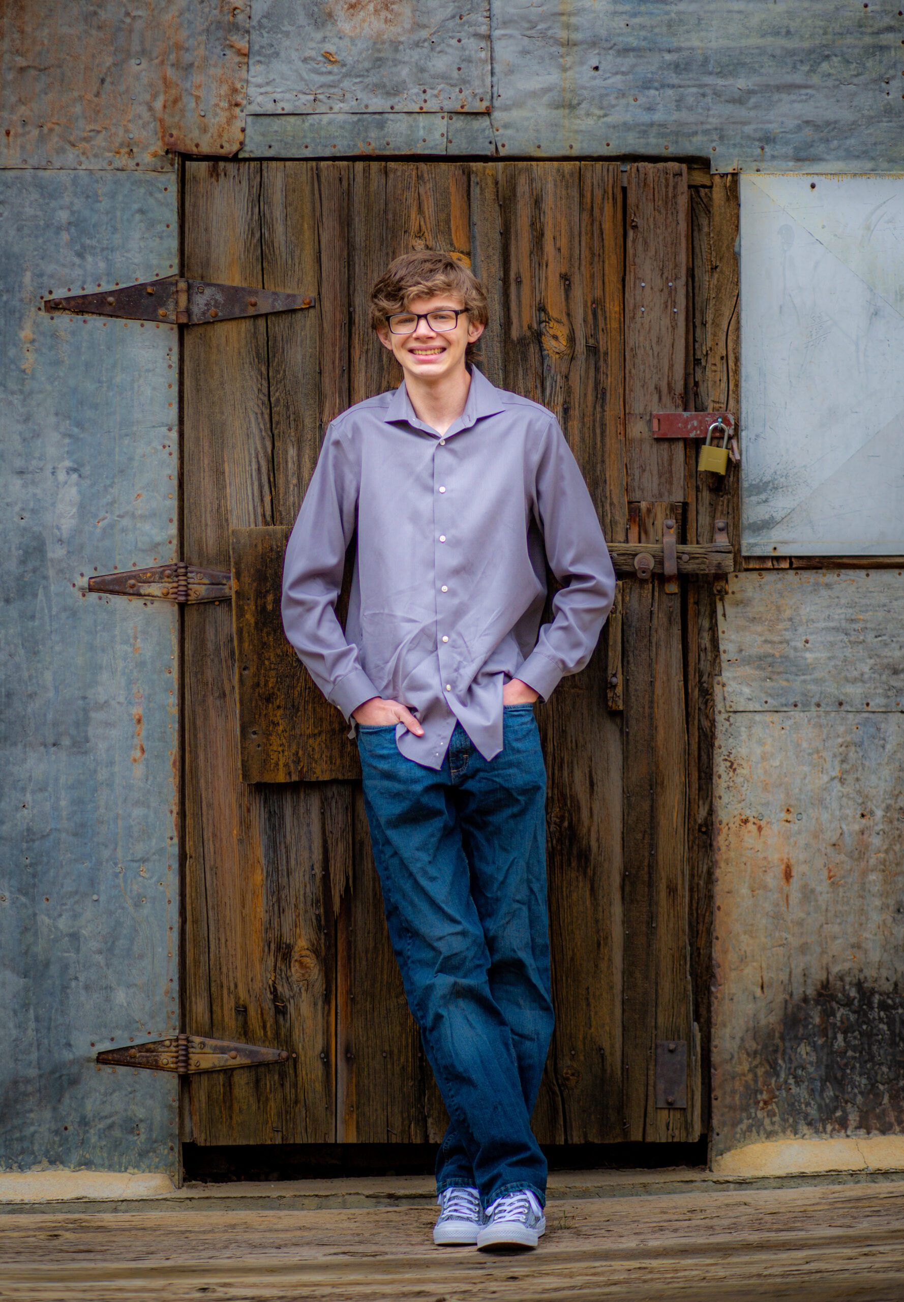 Cheerful young man with glasses wearing a lavender shirt and blue jeans in front of an old textured wooden door