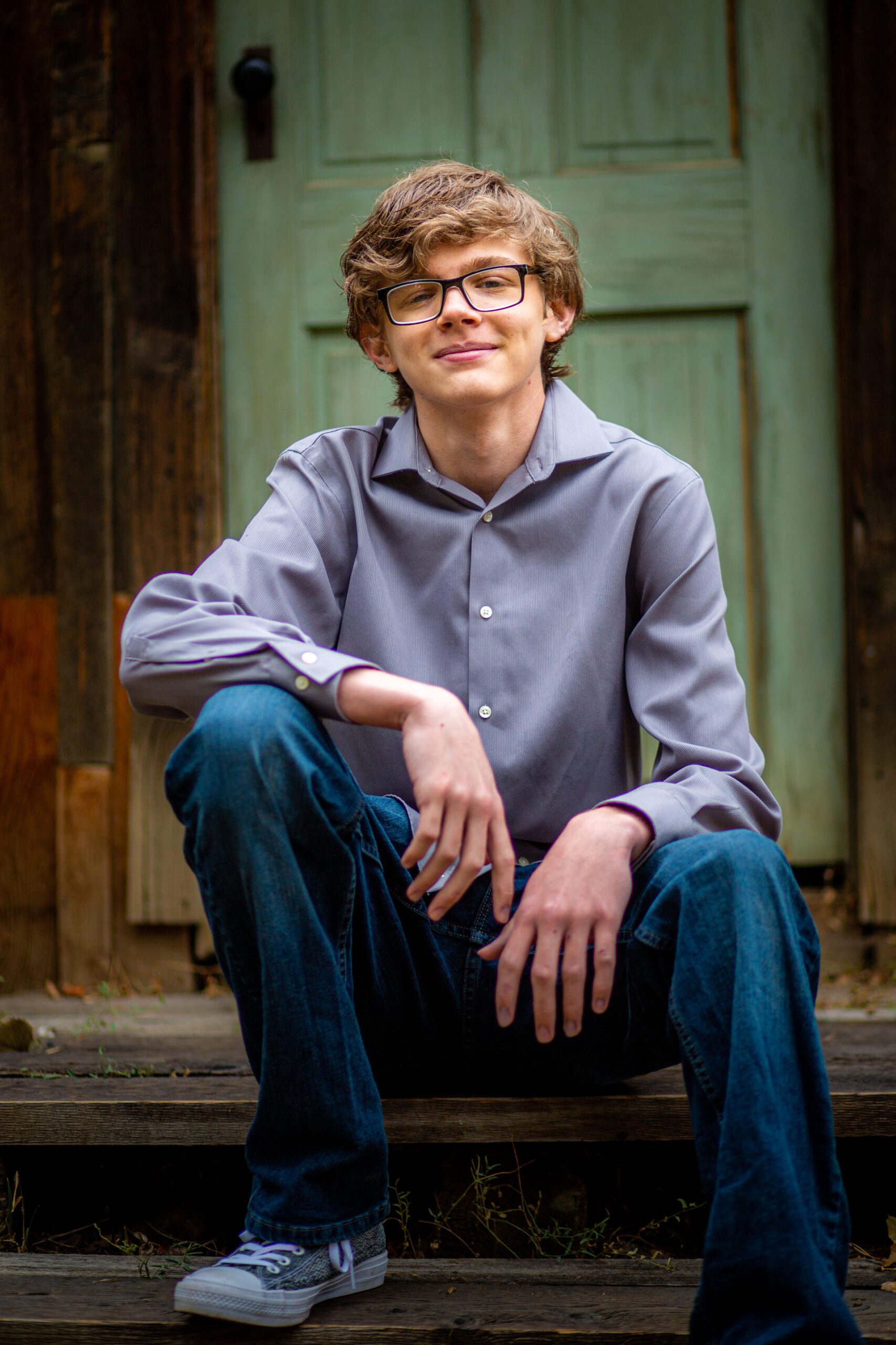 Confident senior man with curly hair and trendy glasses sitting on old wooden steps, emanating a relaxed and studious vibe