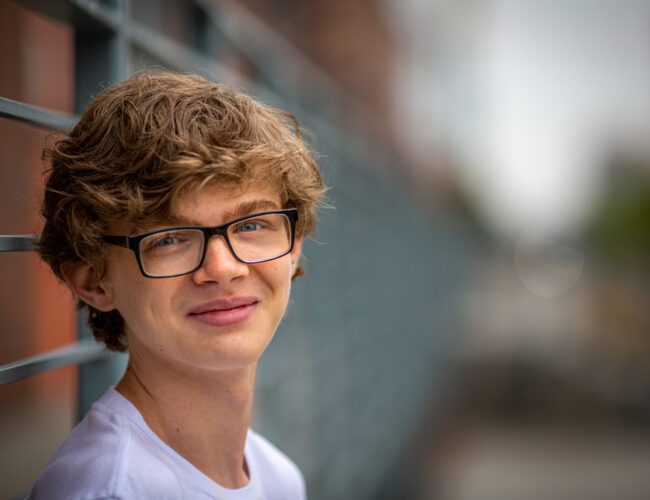 Smiling curly-haired senior man with glasses leaning on a metal railing with an out-of-focus urban background