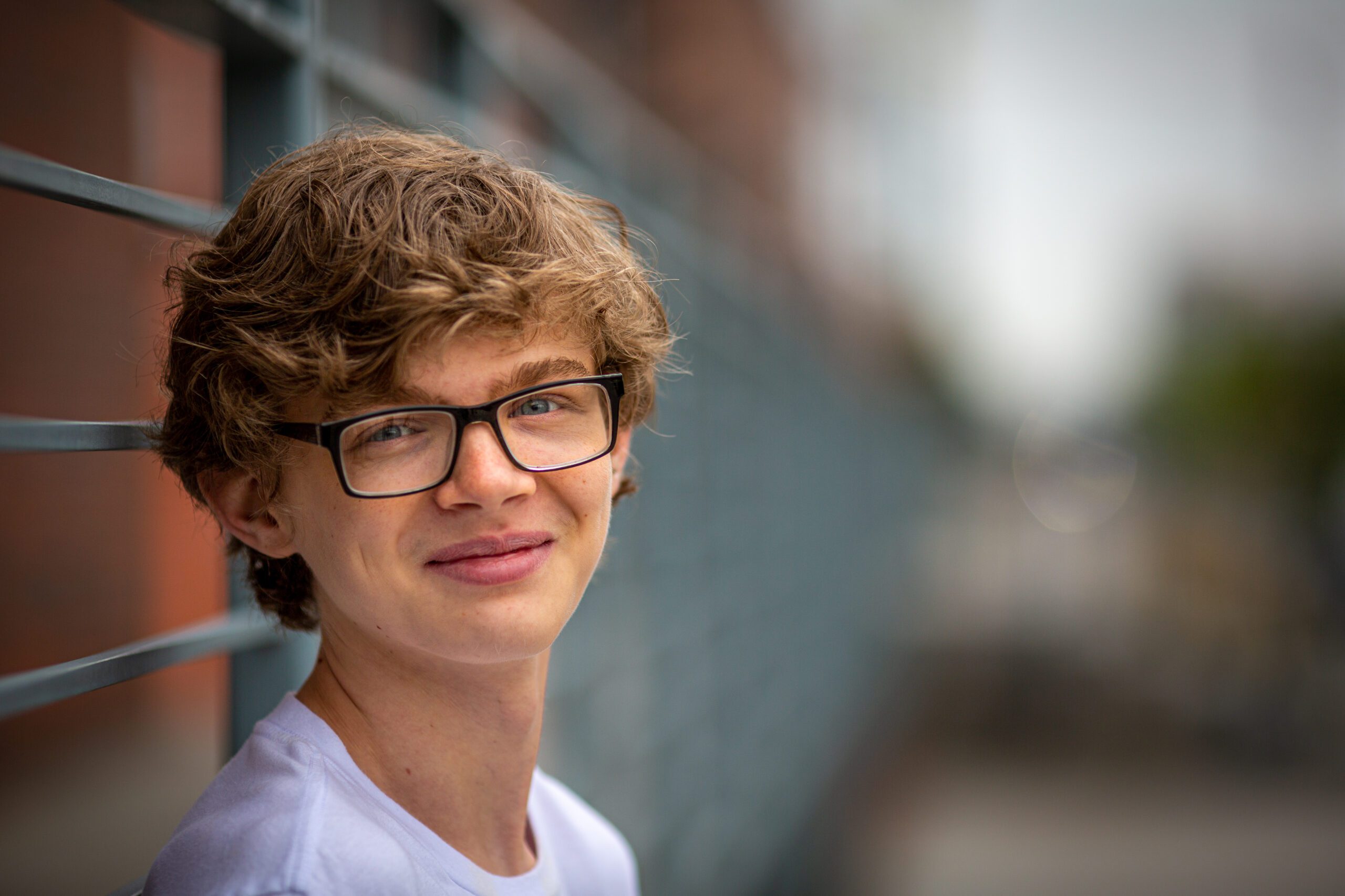 Smiling curly-haired senior man with glasses leaning on a metal railing with an out-of-focus urban background