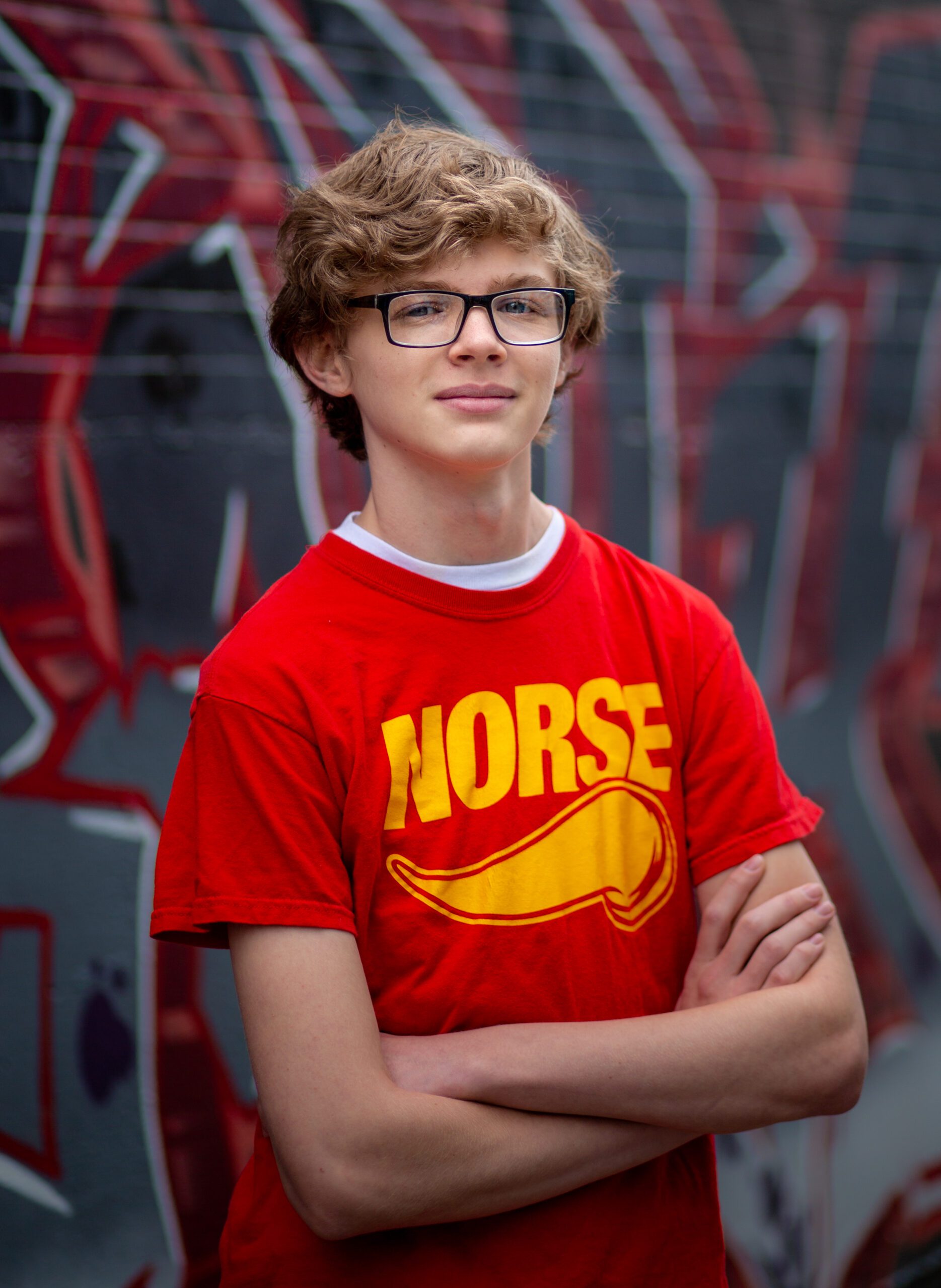 Smiling senior with curly hair and glasses, arms crossed, wearing a red 'NORSE' t-shirt against a graffiti background