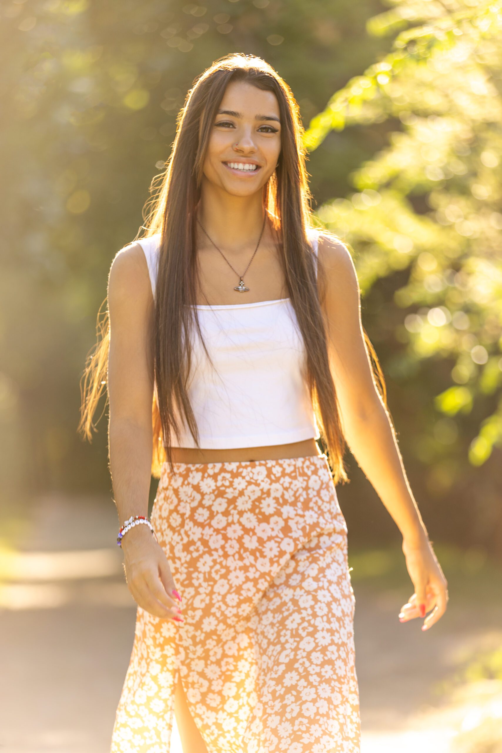 Smiling senior woman with straight long hair wearing a white crop top and floral skirt bathed in warm sunlight