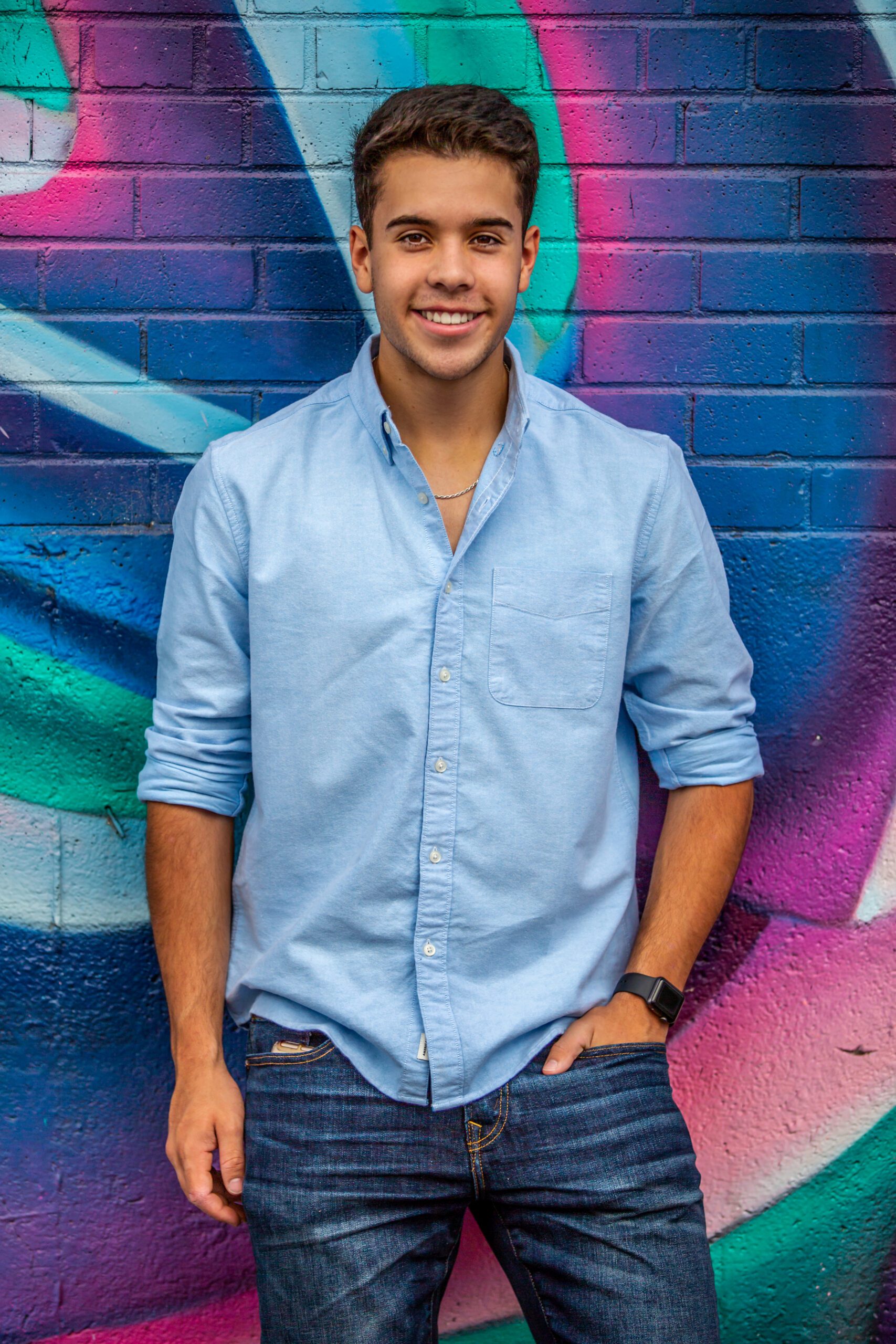 Smiling senior man in a light blue shirt and denim jeans standing against a colorful graffiti background