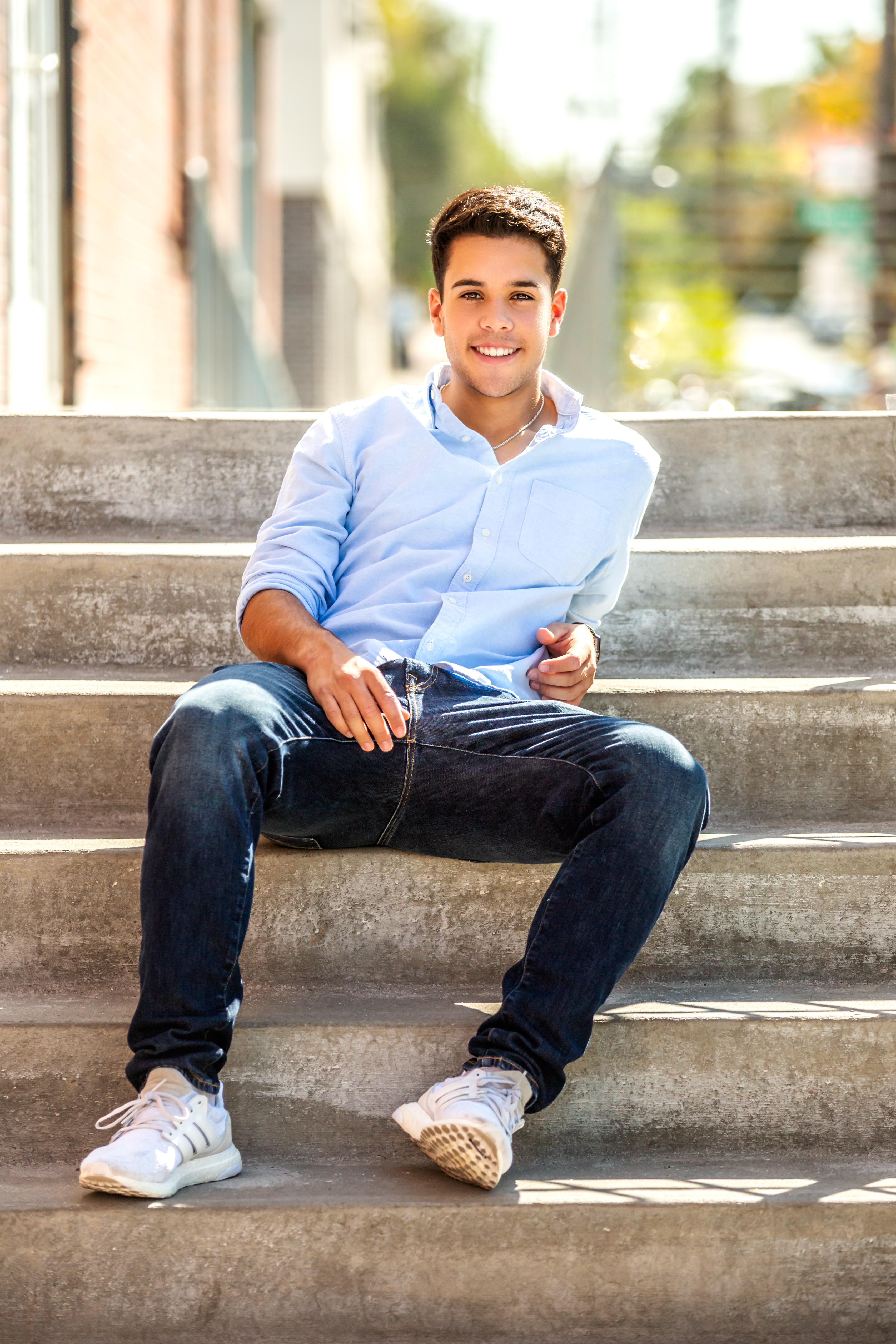 Smiling senior man in a light blue shirt and dark jeans sitting on steps outdoors, holding sunglasses