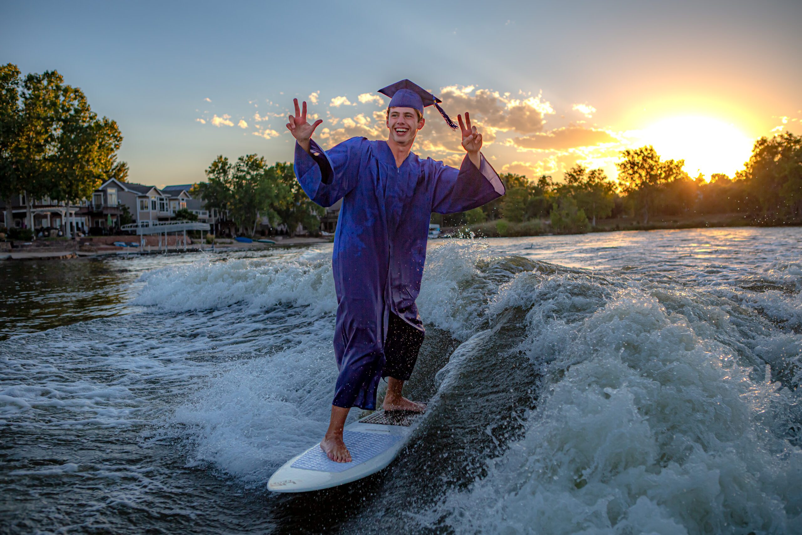 Joyful senior graduate surfing on a wave in full graduation attire during a vibrant sunset