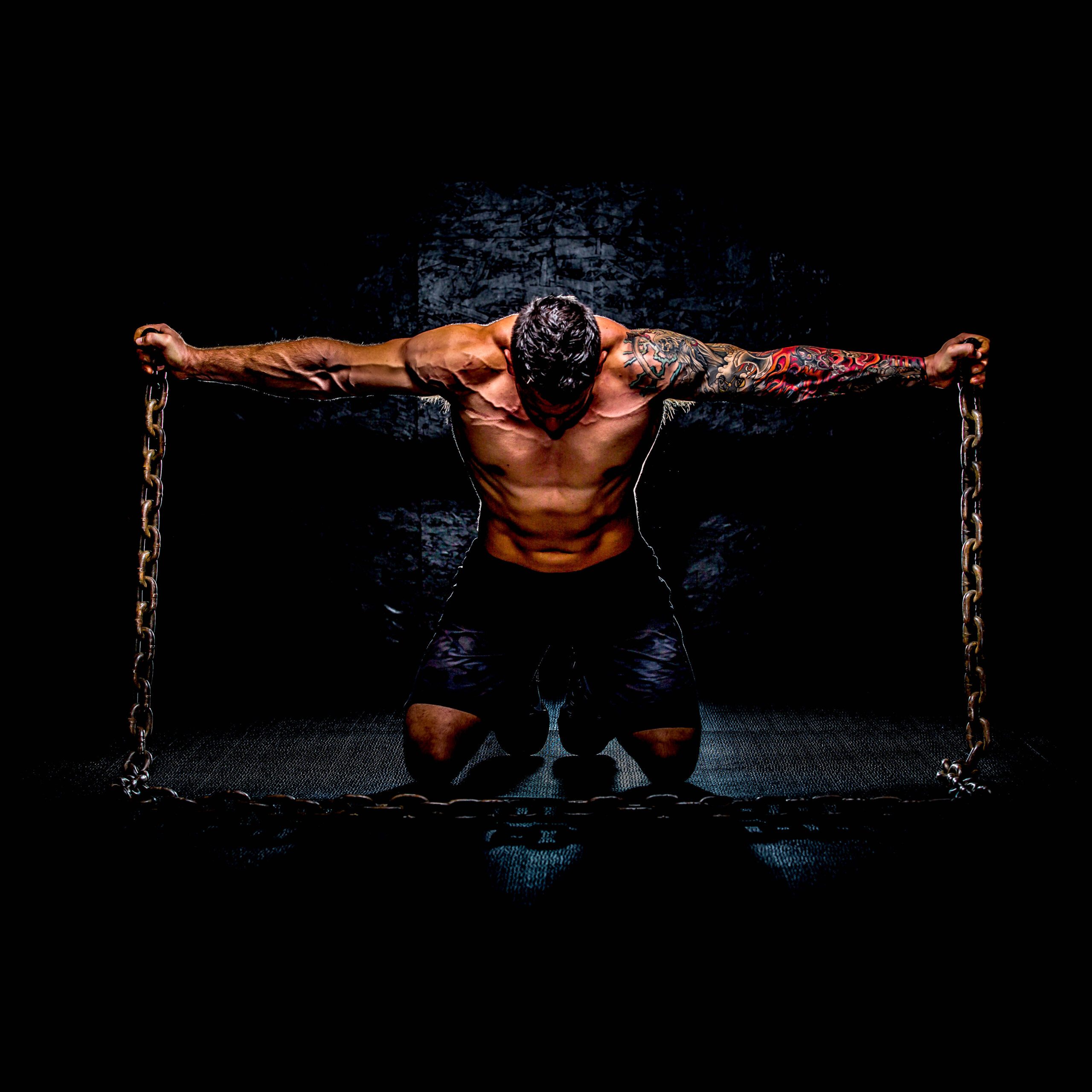 Muscular person with a tattooed arm holding heavy chains in a kneeling pose, with dramatic lighting against a dark background