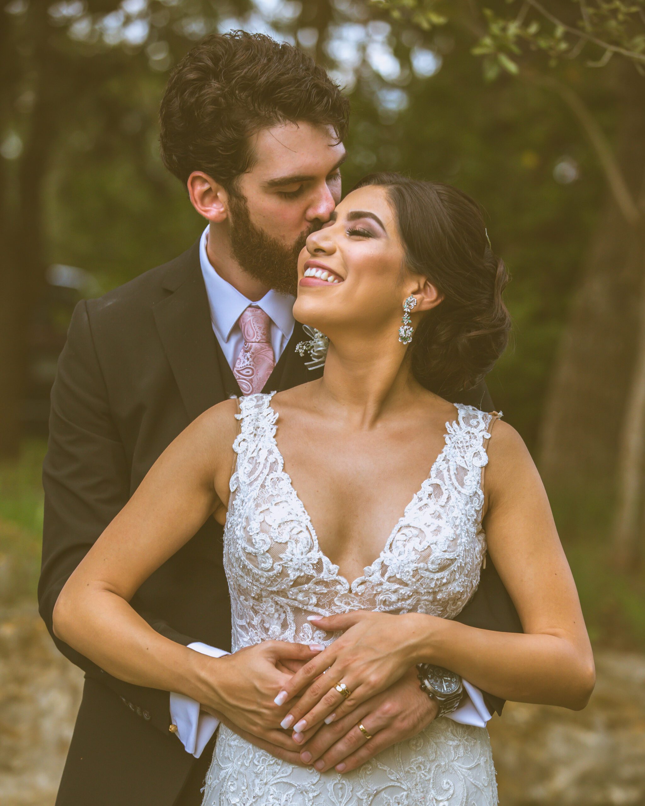 Groom kissing bride on the forehead, both dressed in wedding attire, with a natural backdrop