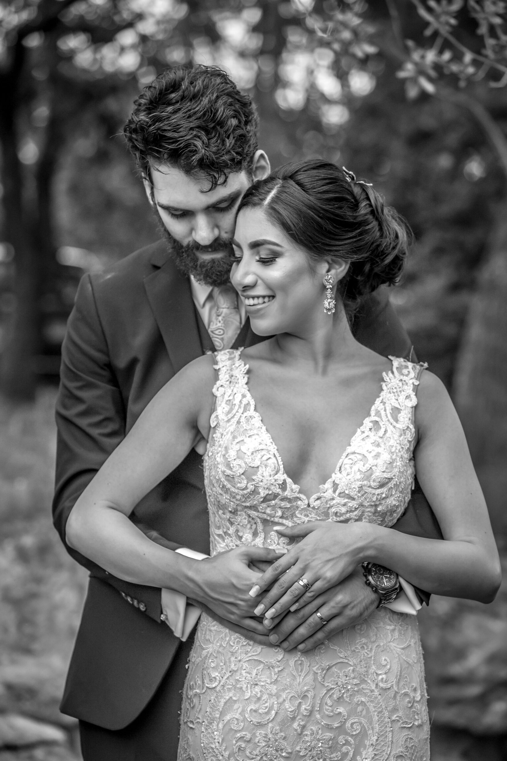 Newlywed couple in an intimate embrace, with the groom in a dark suit and the bride in a lace gown, in a black and white photo