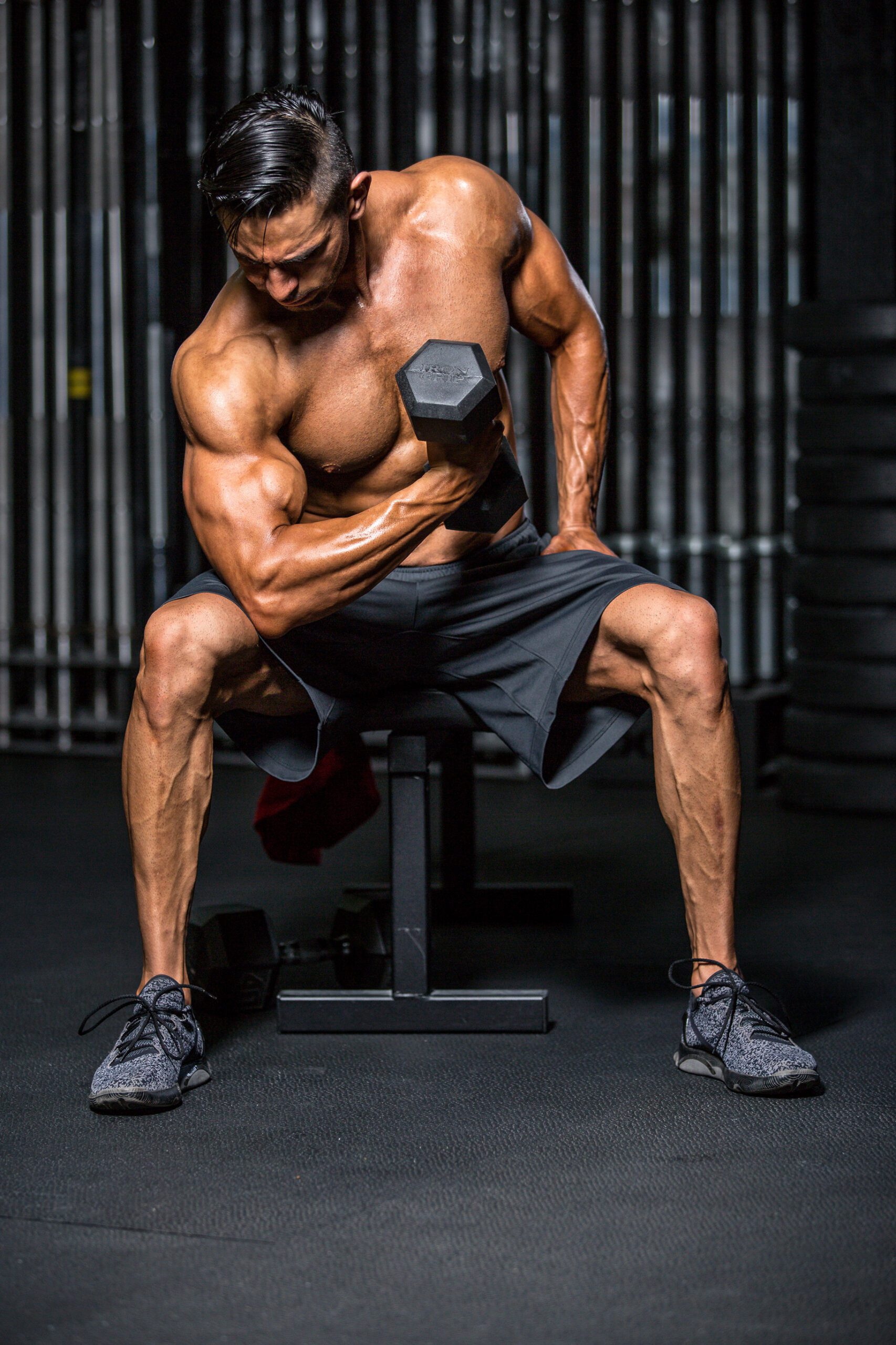 Fit male bodybuilder lifts a dumbbell while sitting on a bench in a gym.