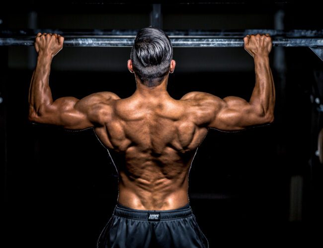 Bodybuilder performing a pull-up on a black and chalky bar with dynamic lighting to highlight his back muscles.