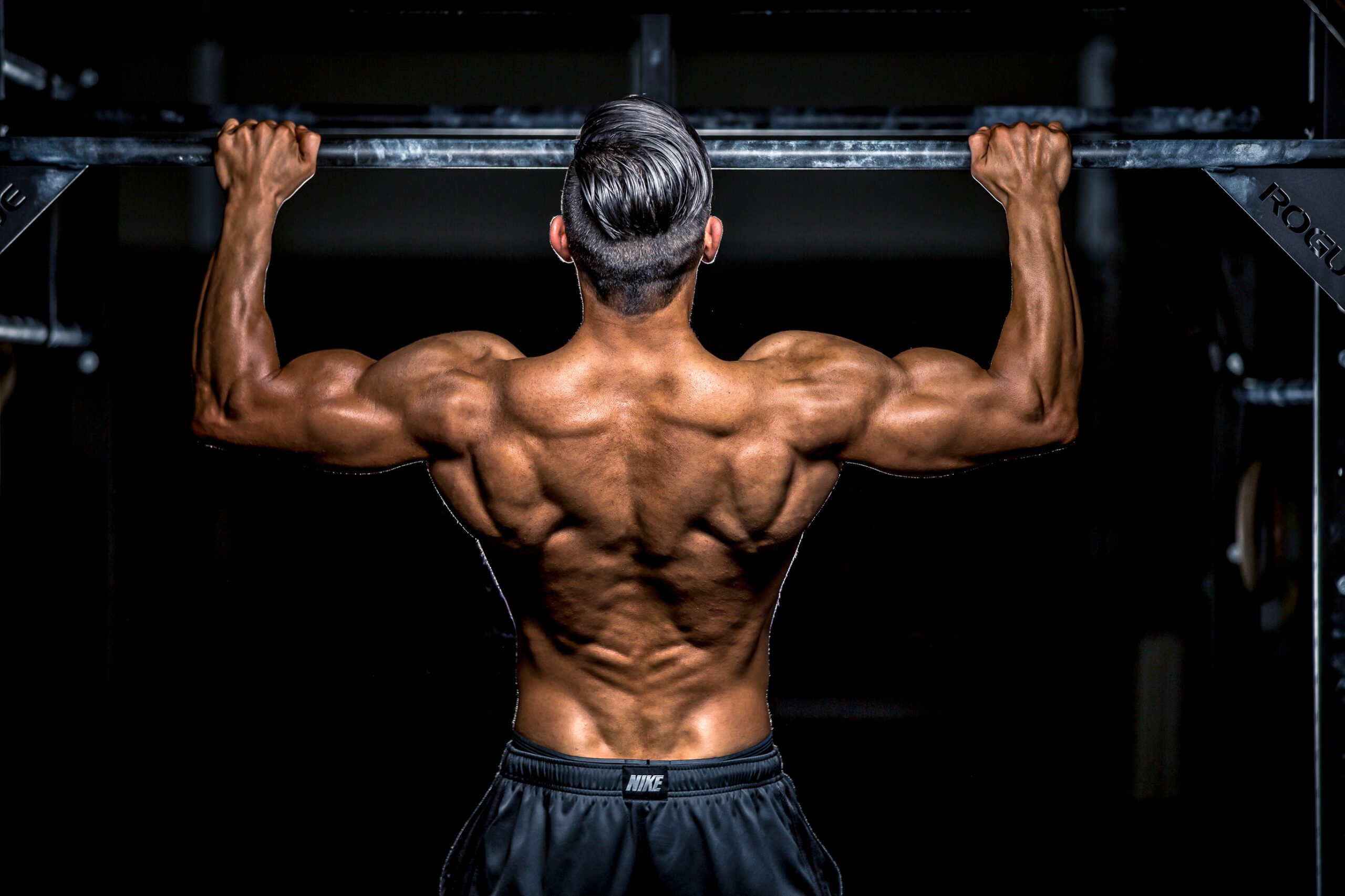 Bodybuilder performing a pull-up on a black and chalky bar with dynamic lighting to highlight his back muscles.