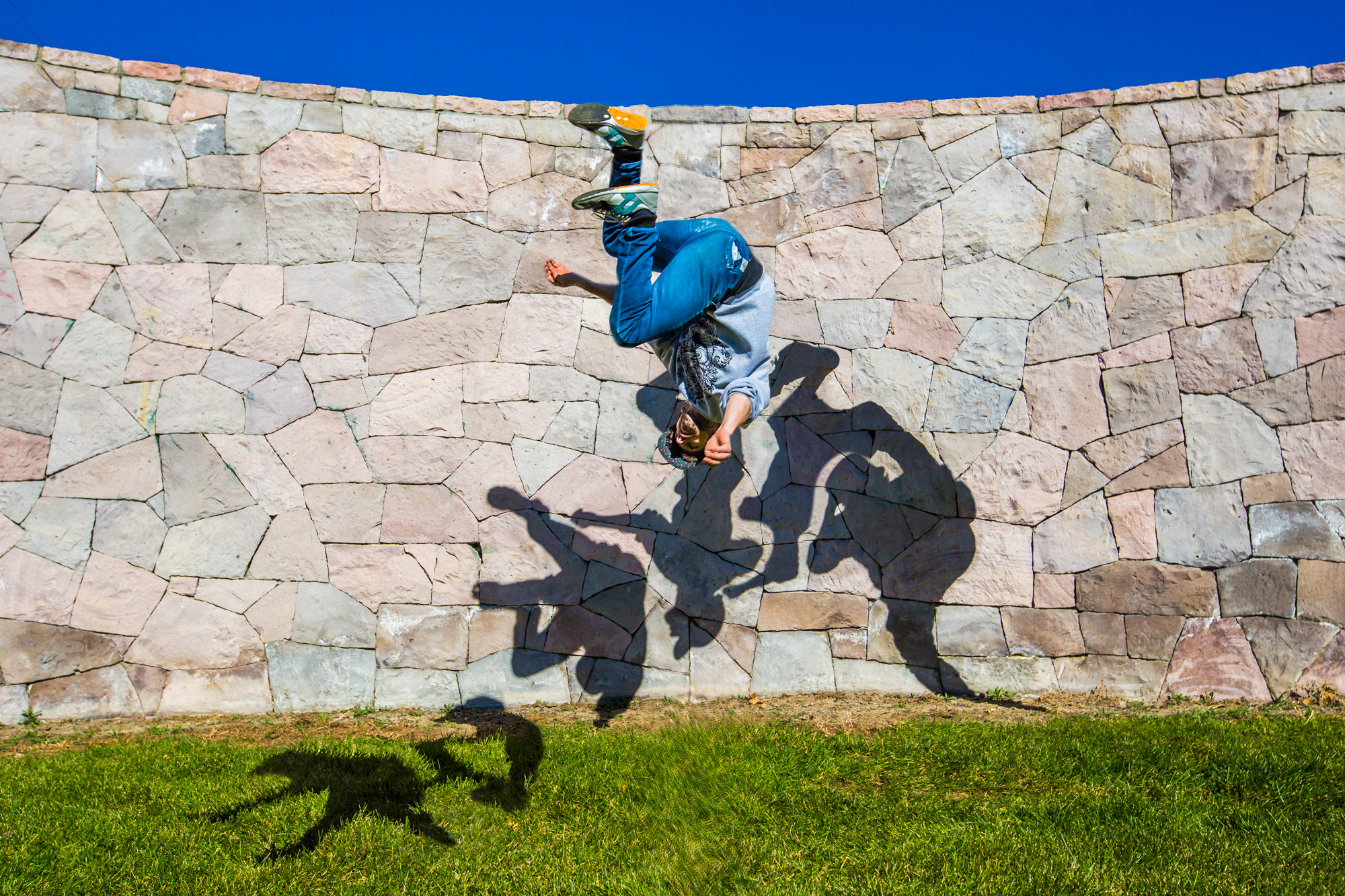 Athletic person doing a backflip against a curved, multicolored stone wall with a detailed shadow mirroring the action, set against a vibrant blue sky